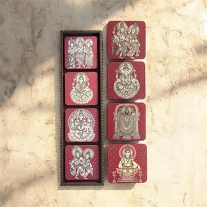 Pode incluir: Um conjunto de oito bases quadradas com figuras religiosas brancas sobre um fundo vermelho. As bases s&atilde;o apresentadas num suporte de madeira castanha. Cada base apresenta uma divindade ou cena diferente, com desenhos detalhados a tra&ccedil;o.