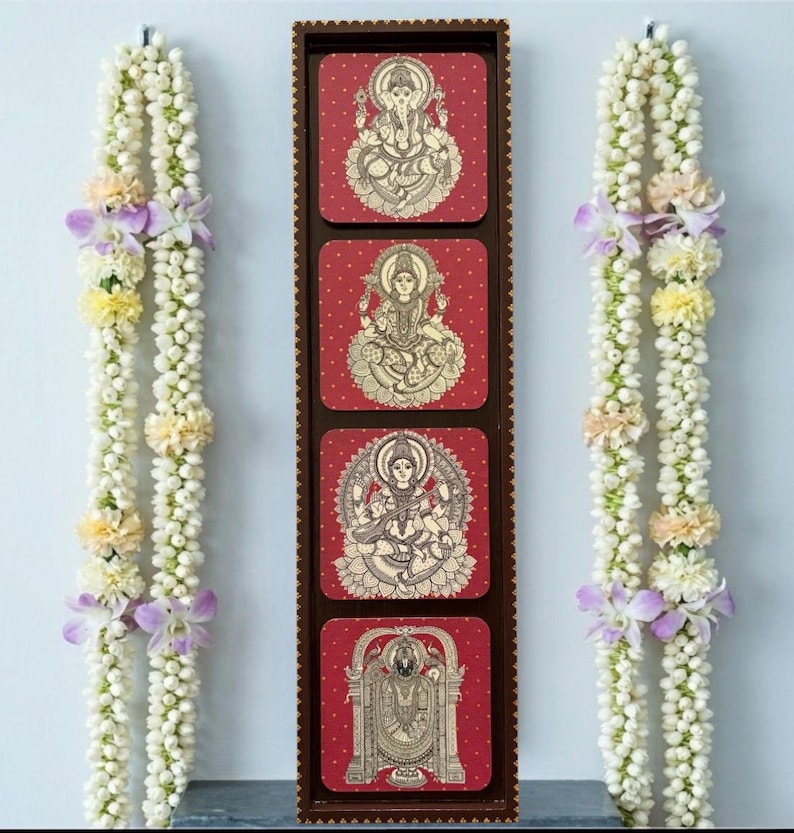 Pode incluir: Uma moldura vertical de madeira com quatro pain&eacute;is quadrados vermelhos, cada um com uma ilustra&ccedil;&atilde;o de uma divindade hindu em bege. Duas guirlandas de flores brancas, com detalhes em roxo e amarelo, ladeiam a moldura.