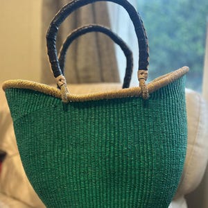 May include: A green woven basket with a rounded shape and two arched handles. The handles are dark brown and the rim is trimmed with a lighter brown material. The basket is on a light-coloured surface.