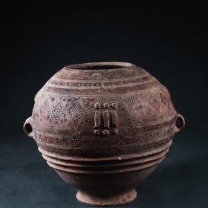 May include: A large, round, terracotta pot with two handles and intricate carved designs. The pot has a wide base and a narrow opening.