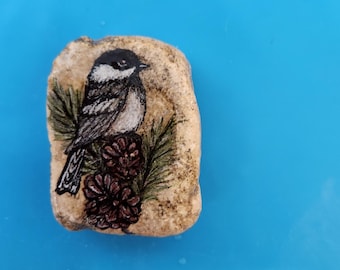 May include: A hand-painted stone features a black-capped chickadee perched on a pine branch with two pine cones. The bird has a black head, white cheeks, and a gray and black body. The stone is beige and has a natural, textured surface.