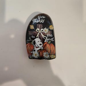 Może przedstawiać: Ręcznie malowany kamień przedstawiający Snoopy'ego, dynie i napis "give thanks to the Lord". Grafika zawiera krzyż, kwiaty i jesienne liście, wszystko na czarnym tle. Uroczy, dekoracyjny element.