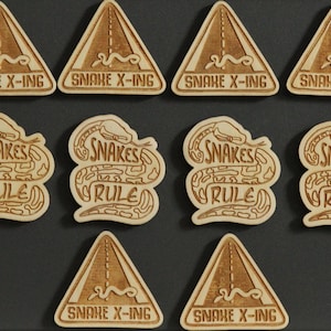 May include: Ten wooden laser-cut signs with a snake theme.  Four signs feature a snake crossing road sign with the text "SNAKE X-ING". Six signs feature a snake wrapped around the text "SNAKES RULE".