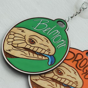May include: Two circular wooden ornaments featuring laser-engraved lizard heads with blue tongues and orange eyes. The top ornament is green with the text Belmont, and the bottom is orange with the text Drogo. Both have silver chains and suction cups.