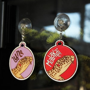 Puede incluir: Dos etiquetas redondas de madera para mascotas con forma de gecko, con ventosas. La etiqueta rosa dice "Luna" y la etiqueta roja dice "Malakai".