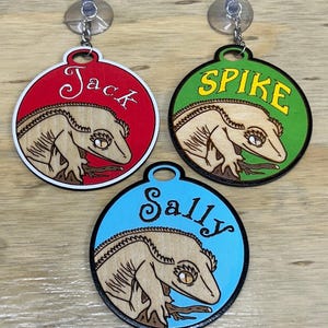 May include: Three circular wooden ornaments with suction cups, each featuring a laser-engraved crested gecko on a branch. The red ornament says Jack, the green one says SPIKE, and the blue one says Sally in distinct fonts.