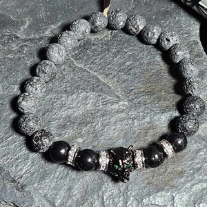 May include: Black lava rock beaded bracelet with a black panther charm and silver accents. The panther has green eyes.