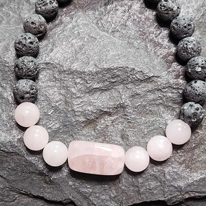 May include: A bracelet made with black lava rock beads and pink rose quartz beads. The bracelet has a larger rectangular rose quartz bead in the center.