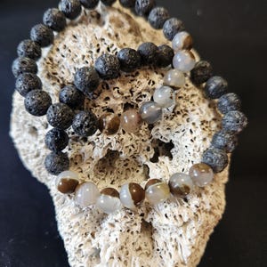 May include: Two bracelets, one made of black lava rock beads and the other made of brown and white agate beads.