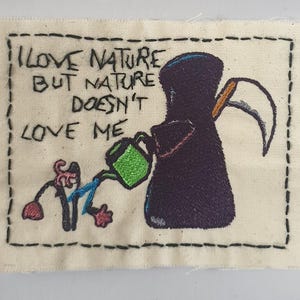 Könnte beinhalten: Gestickter Aufnäher mit dem Text "I LOVE NATURE BUT NATURE DOESN'T LOVE ME". Der Aufnäher zeigt eine Sensenmann-Figur, die eine kleine Figur mit einer Gießkanne gießt. Der Aufnäher befindet sich auf einem cremefarbenen Stoff mit schwarzen Nähten.