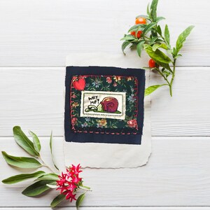 Puede incluir: Una pieza de arte textil hecha a mano con una ilustración de un caracol y el texto "WHY ME?" sobre un fondo floral. La obra de arte está enmarcada con un borde azul oscuro y un corazón rojo. Hojas verdes y flores rojas están dispuestas alrededor.