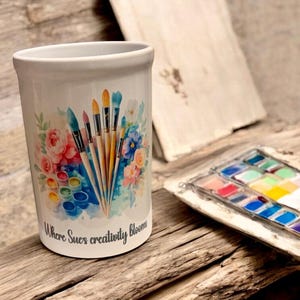 May include: White ceramic brush holder featuring a watercolour design of paintbrushes, flowers, and paint palettes. The text "Where Sues creativity blooms" is printed on the holder. A watercolour paint set is visible.