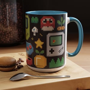 May include: A white ceramic mug with a blue interior and handle. The mug has a black background with colourful pixelated graphics of retro video game elements. A macaron, coffee beans, and a spoon are on a wooden surface.