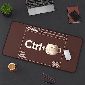 Puede incluir: Una alfombrilla de escritorio marrón con el texto "Coffee Ctrl+C Brew Copy Repeat". El diseño incluye un gráfico de taza de café y la frase "Your daily dose of motivation, one sip at a time". También se ven un ratón blanco y un portátil.