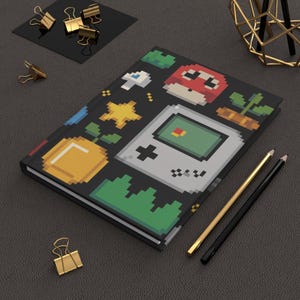 Puede incluir: Un diario negro con un diseño de videojuego pixelado que presenta un hongo, una estrella y una consola portátil. Lápices dorados y negros y clips dorados están sobre la superficie gris oscuro.
