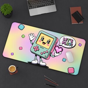 May include: A rectangular mousepad with a pastel rainbow gradient background. A cartoon handheld game console character is featured, holding a pixelated heart and a speech bubble that says "LET'S PLAY!". Colorful geometric shapes are scattered around.