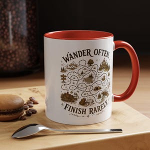 May include: A white ceramic mug with a bright red interior and handle. The exterior features a brown ink illustration of a winding path through a landscape with mountains, a castle, and a house, accompanied by the text Wander Often, Finish Rarely.