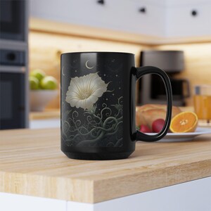 May include: Black ceramic mug featuring a white flower design with a detailed stem and leaf pattern. The design includes a crescent moon and stars on a dark background. The mug has a large handle and is suitable for hot beverages.