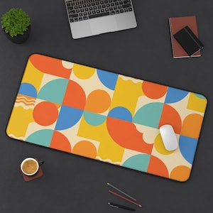 May include: A rectangular desk mat with a colorful geometric pattern. The design features orange, yellow, blue, and cream-colored shapes. A white computer mouse rests on the mat, alongside a laptop, coffee cup, and stationery.