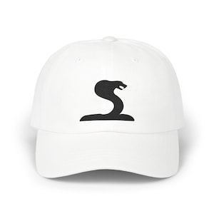 May include: A white baseball cap featuring a black embroidered graphic of a cobra snake in a coiled, upright position on the front panel. The cap has a structured crown, a curved brim, and a button on the top centre of the dome-shaped headwear.
