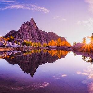 May include: A panoramic view of a mountain lake with a sharp, rocky peak reflected in the still water. The sun is setting behind the mountain, casting a golden glow on the trees and the water.