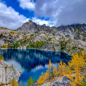 May include: A scenic view of a mountain lake surrounded by rocky cliffs and lush green trees. The water is a deep blue and reflects the clouds in the sky. The trees in the foreground are a vibrant yellow, adding a pop of color to the scene.