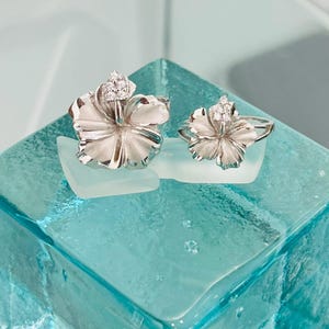 Może przedstawiać: Dwa srebrne pierścionki z białym wzorem kwiatu i przezroczystymi kamieniami. Pierścionki leżą na niebieskiej szklanej powierzchni.