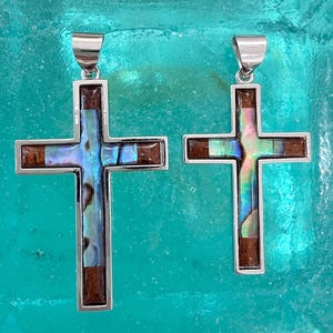 Può includere: Due ciondoli a croce in argento con inserti in legno marrone e conchiglia di abalone iridescente. Ogni croce presenta una cornice in argento lucido e un gancio in argento sulla parte superiore. La croce più grande è a sinistra, la piccola a destra.