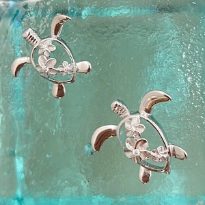 May include: A pair of silver earrings featuring a detailed turtle design with white flower accents.