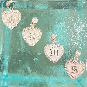 Puede incluir: Cuatro colgantes de plata en forma de corazón con diferentes letras grabadas. Las letras son C, K, M y S. Los colgantes están sobre un fondo azul claro.