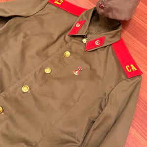 Peut inclure: Une veste de style militaire marron avec des épaulettes rouges portant les lettres jaunes CA, des pattes de col rouges avec des emblèmes circulaires dorés, des boutons dorés et une petite médaille. Une toque marron assortie avec une étoile rouge.