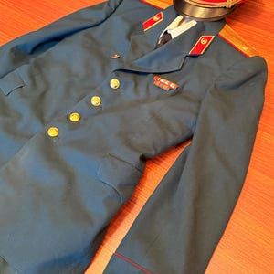 Peut inclure: Une veste de cérémonie de style militaire bleu sarcelle foncé avec des boutons dorés, des pattes de col rouges à emblèmes dorés et des épaulettes. Une casquette assortie à bandeau rouge, jugulaire dorée et étoile centrale est posée au-dessus.
