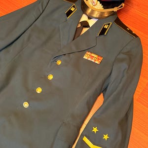 Könnte beinhalten: Eine dunkel-petrolfarbene Jacke im Militärstil mit goldfarbenen Knöpfen, schwarzen Kragenspiegeln mit goldenen Abzeichen sowie gelben Stern- und Winkelaufnähern am Ärmel. Dazu eine passende Schirmmütze mit rotem Band und Sternemblem.