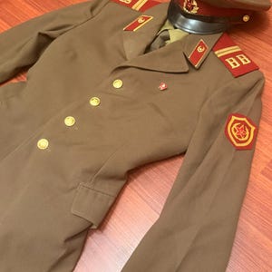 May include: A tan military-style jacket with gold-tone buttons, red collar tabs, and shoulder boards featuring the letters BB. A matching tan peaked cap with a red band and a central emblem featuring a red star sits atop the jacket collar.