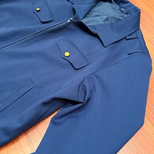 Könnte beinhalten: Eine einfarbige marineblaue Jacke mit durchgehendem Frontreißverschluss, spitzem Kragen und zwei Brusttaschen mit spitzen Klappen, die mit kleinen goldfarbenen Knöpfen verschlossen werden. Die Jacke hat lange Ärmel und ein dunkles Innenfutter.