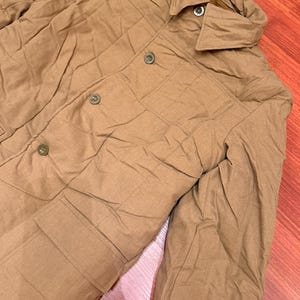 May include: A tan quilted jacket featuring a pointed collar, a front button closure with four visible buttons, and two large patch pockets with flaps on the lower front. The garment has long sleeves and a textured, slightly wrinkled fabric finish.
