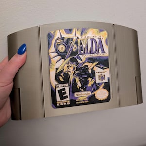 May include: A Nintendo 64 video game cartridge for "The Legend of Zelda: Majora's Mask." The cartridge has a gray plastic shell with a colorful label featuring game art and the ESRB rating. The label includes the Nintendo logo.