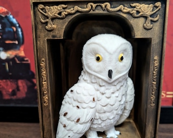 Könnte beinhalten: Eine weiße Schneeeulenfigur mit gelben Augen wird in einem goldfarbenen Rahmen mit verzierten Details präsentiert. Die Eule sitzt auf einer kleinen Plattform im Rahmen.
