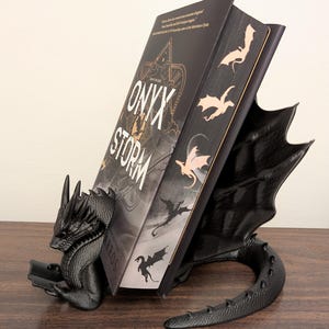 May include: Black dragon bookend holding a book titled "Onyx & Storm" by Rosaria Munda. The bookend is made of 3D printed plastic and features a detailed dragon design.