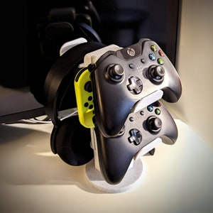 Peut inclure: Un support de casque de jeu noir et gris avec trois emplacements pour manettes. Le support tient deux manettes Xbox noires et une manette Nintendo Switch verte et jaune.