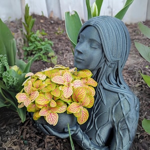 May include: A gray resin sculpture of a woman with long hair, holding a pot of yellow and red foliage. The sculpture is placed in a garden setting.