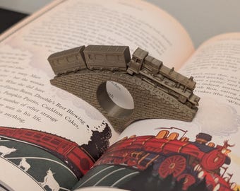May include: A miniature train engine and boxcars crossing a bridge made of gray metal. The train is on top of a book with a red cover and illustrations of a train and cows. The book's text reads "Drooble's Best Blowing Cauldron Cakes."