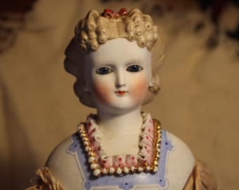 May include: A porcelain doll with a white face, blue eyes, and pink cheeks. The doll is wearing a white dress with a blue and gold trim and a pink and white beaded necklace.