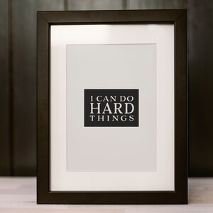 May include: A black picture frame with a white mat and a white background. The frame contains a black and white print with the text "I can do hard things".