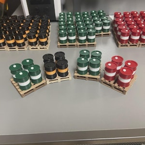 Może przedstawiać: Miniaturowe beczki w kolorze czarnym, pomarańczowym, zielonym i czerwonym, z białymi pasami, ułożone są na małych drewnianych paletach. Beczki są pogrupowane w różnych ilościach, co sugeruje koncepcję ekspozycji lub przechowywania produktów.