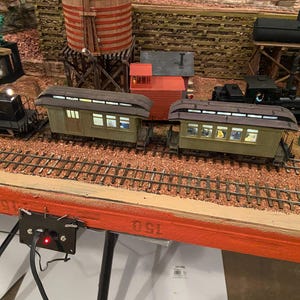 Puede incluir: Un detallado juego de trenes modelo con una locomotora negra, vagones de pasajeros y un vagón de cola rojo. La escena incluye una torre de agua, edificios y vías férreas en miniatura, todo sobre una base de diorama. El modelo está iluminado.
