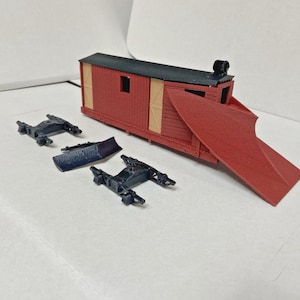 Puede incluir: Un modelo de quitanieves de tren con cabina roja y techo negro, y una cuchilla roja. También se incluyen dos bogies negros y una cuchilla azul oscuro. El modelo está sobre una superficie blanca.