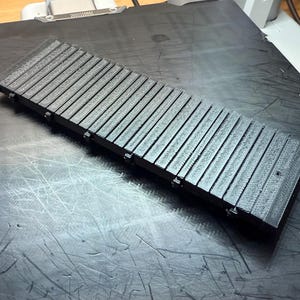 May include: A rectangular, black, 3D-printed object with a series of parallel, ridged slats. The object is lying on a dark surface. The slats are evenly spaced and run the length of the object.