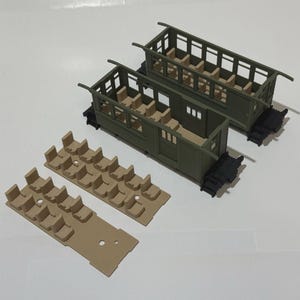 Può includere: Sono esposti due modelli di vagoni ferroviari verde oliva con sedili beige e carrelli neri. Sono presenti anche due inserti per sedili beige. I vagoni hanno fianchi e tetti aperti, con sedili interni dettagliati visibili.