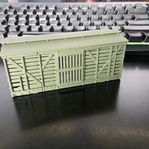 Puede incluir: Un vagón de tren modelo verde salvia con un diseño de madera de listones. El vagón tiene un techo acanalado y una puerta corredera. El modelo está sobre un fondo de teclado negro.